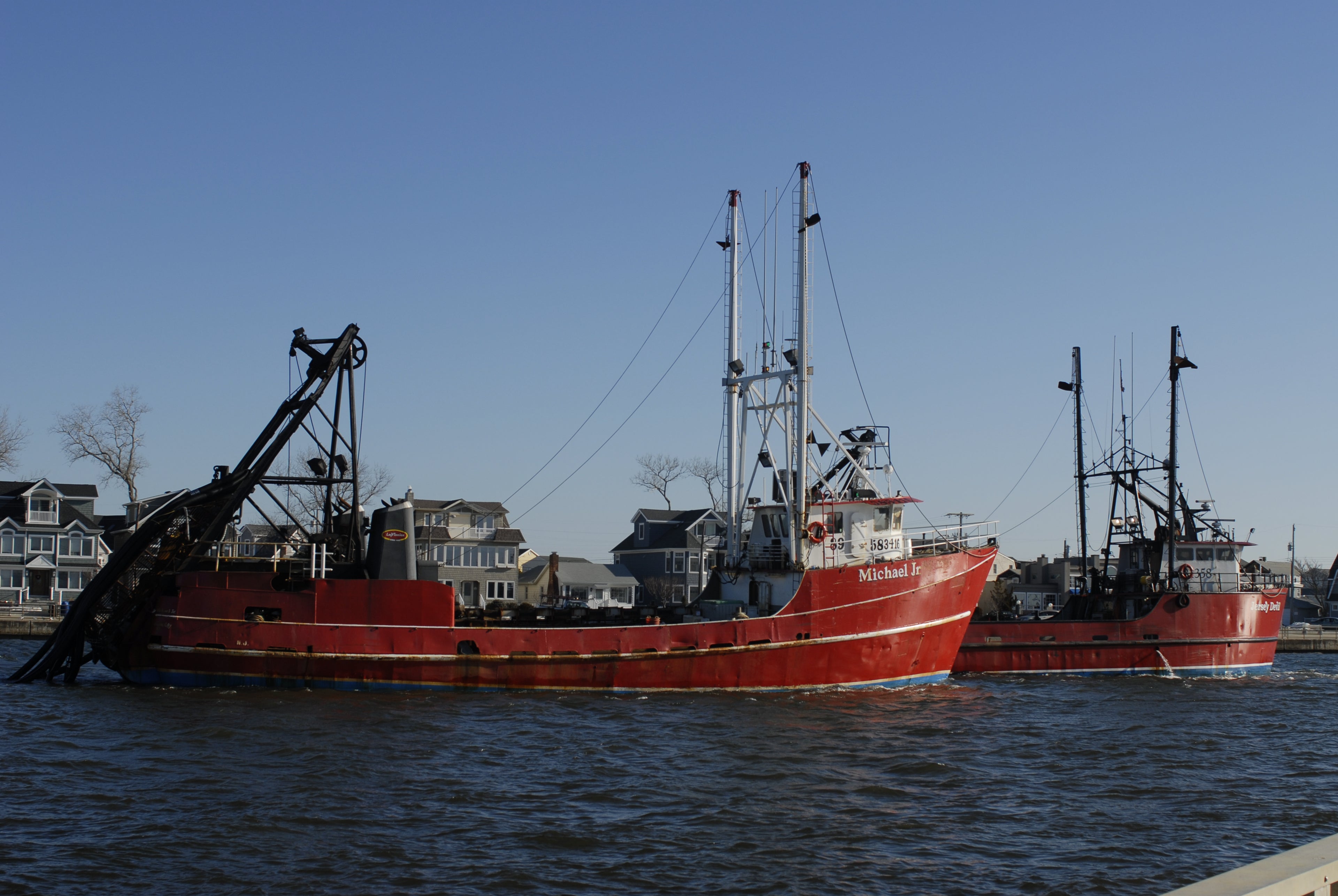 Two large red fishing boats on a body of water with houses in the background.