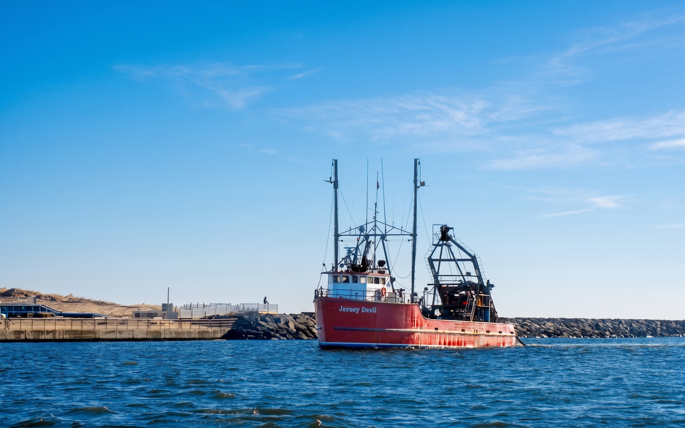 A red fishing boat with a white cabin and tall masts floats on blue water under a clear blue sky, with a rocky breakwater and low shoreline in the background.