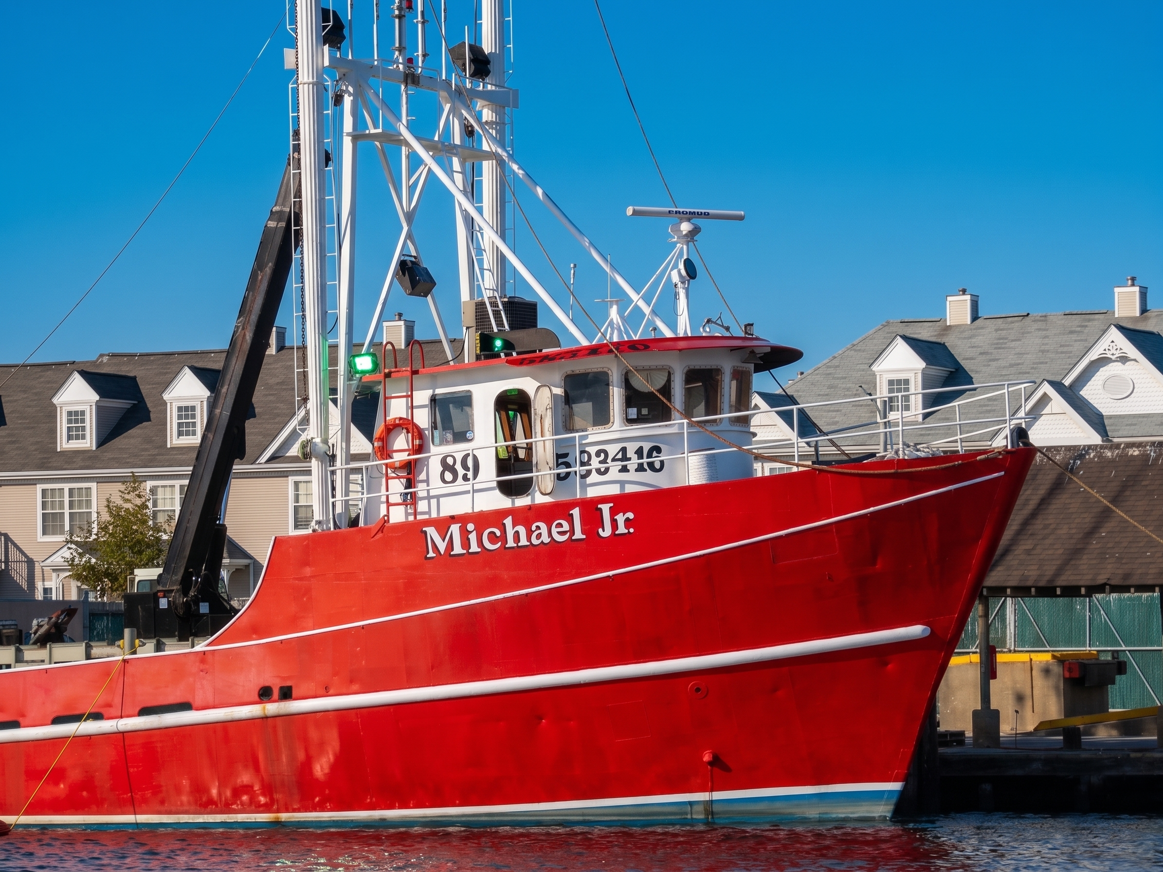 Red fishing boat named 'Michael Jr' docked at a harbor with houses in the background.